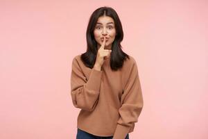 Indoor photo of young agitated long haired brunette lady raising hand with hush gesture while asking to keep silence, isolated over pink background in casual wear