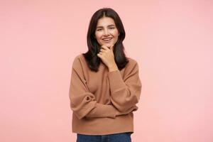 Pleased young lovely brown haired female with loose hair holding her chin with raised hand and looking gladly at camera with nice smile, isolated over pink background photo