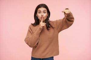 Excited young brown haired lady showing emotionally at camera and keeping forefinger on her mouth while asking to keep silence, posing over pink background photo