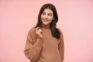 Cheerful young charming brunette female with natural makeup touching her hair with raised hand and looking gladly aside, standing over pink background photo