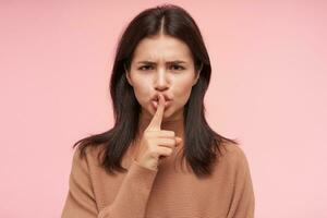 Severe young brunette female with natural makeup frowning her eyebrows while looking seriously at camera, asking to be quiet and holding forefinger on her lips, isolated over pink background photo