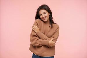 Cheerful young attractive brunette woman dressed in knitted poloneck emracing herself and looking at camera with pleasant smile, isolated over pink background photo