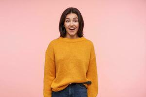 Excited young lovely brown haired lady keeping her mouth wide opened while looking emotionally at camera, standing over pink background in yellow knitted sweater photo