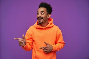 Joyful young attractive bearded dark skinned curly brunette male raising forefingers and looking aside with wide cheerful smile, dressed in orange hoodie over purple background photo