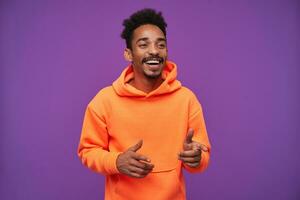 Cheerful young pretty bearded dark skinned man with curly black hair laughing happily and showing aside with raised forefingers, standing against purple background in casual sporty wear photo