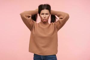 Indoor photo of young offended brunette woman with natural makeup holding ponytails with raised hands and showing sadly her tongue to camera, isolated over pink background