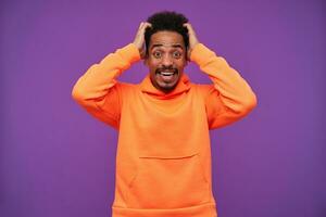 Overwhelmed young dark haired bearded dark skinned guy rounding his brown eyes while looking shocked at camera, holding his head with raised hands while posing over purple background photo