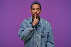 Portrait of young nervous dark skinned brunette man with beard frowning his eyebrows while looking at camera and raising hand in hush gesture while standing over purple background photo