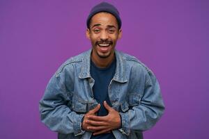 Indoor photo of young attractive dark skinned brunette male with beard cluthing at his belly with hands while laughing joyfully while looking happily at camera, isolated over purple background
