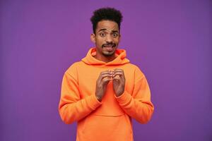 Puzzled young curly dark haired bearded man with dark skin frowning confusedly his face and folding raised hands while posing over purple background, dressed in casual orange hoodie photo