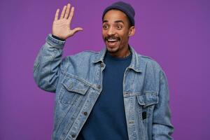 Happy young pretty dark skinned bearded brunette male rojoicing while meeting friend and raising palm in hello gesture, smiling happily while posing over purple background photo