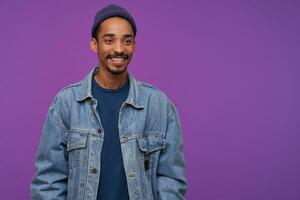 Cheerful young attractive bearded brunette man with dark skin looking positively aside with charming smile, wearing blue cap, pullover and jeans coat while posing over purple background photo