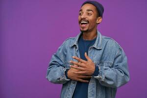 Joyous young handsome dark skinned bearded brunette male smiling joyfully and looking happily aside, dressed in casual clothes while posing over purple background photo