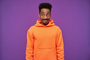 Studio shot of young lovely bearded dark skinned curly brunette guy looking positively aside with confused grimace, keeping hands down while standing over purple background photo