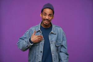 Puzzled young attractive dark skinned bearded brown haired man showing on himself with raised palm while looking confusedly to camera, standing against purple background photo