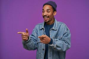 Studio photo of young cheerful lovely dark skinned brunette man with beard pointing aside with raised forefingers and smiling widely while standing over purple background