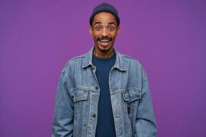 Indoor shot of young joyful dark skinned male with beard looking at camera with excited face, raising happily eyebrows while smiling widely, posing over purple background photo