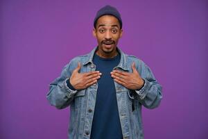 Open-eyed young attractive dark skinned brunette male with beard holding palms on his chest and looking at camera with surprised face, posing over purple background in casual wear photo