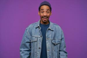 Open-eyed young beautiful dark haired bearded male in blue cap, pullover and jeans coat raising amazedly eyebrows while looking joyfully at camera, standing over purple background photo