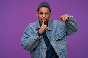 Displeased young handsome dark skinned bearded brunette guy holding index finger on his mouth and frowning his face, asking to keep silence, isolated over purple background photo