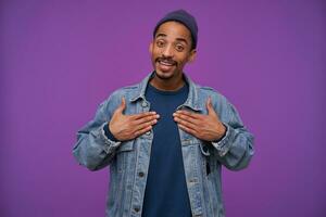 Cheerful young pretty dark skinned bearded guy in blue cap, pullover and jeans coat looking positively at camera with wide sincere smile, standing over purple background with hands on his chest photo