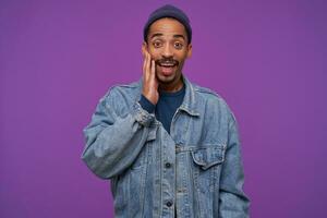 Surprised young pretty brown-eyed dark skinned bearded man keeping raised palm on his cheek while looking wonderingly to camera, standing over purple background photo