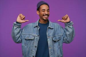 Cheerful young handsome bearded brunette guy with dark skin showing on himself with raised hands and smiling self-confidently at camera, isolated over purple background photo