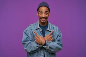 Indoor photo of cheerful young handsome dark skinned bearded man smiling widely while showing with forefingers in different directions, standing over purple background