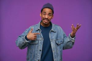Bewildered young dark skinned bearded brunette male wrinkling forehead and raising confusedly hand while looking at camera, wearing casual clothes over purple background photo