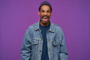 Indoor photo of young angry dark skinned brunette male with beard frowning his face while screaming fiercely with wide mouth opened, isolated over purple background