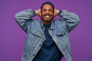 Studio shot of young stressed dark skinned bearded guy frowning his face with closed eyes while covering ears with raised palms, avoiding loud sounds, isolated over purple background photo