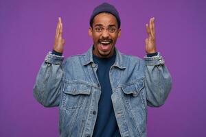 Portrait of young pretty dark skinned bearded guy raising palms emotionally and frowning face while shouting angrily, dressed in casual clothes while posing over purple background photo