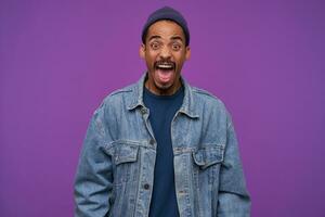 Furious young brown-eyed bearded man with dark skin shouting angrily and wrinkling forehead while looking heatedly at camera, wearing blue cap, pullover and jeans coat over purple background photo