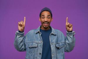 Joyful young handsome dark skinned brunette male with beard showing upwards while standing over purple background, rounding his brown eyes while looking happily at camera photo