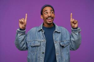 Pleasant looking young pretty bearded brown haired man with dark skin pointing upwards with raised index fingers and smiling positively, standing over purple background photo