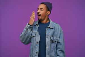 Tired young pretty dark skinned bearded brunette guy covering his mouth with raised palm while yawning with closed eyes, dressed in clothes while posing over purple background photo