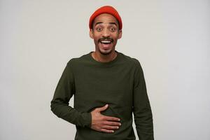 Portrait of overjoyed young bearded dark skinned guy looking at camera with wide eyes opened and holding hand on his belly while laughing happily, isolated over white background photo