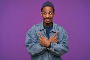 Studio shot of young dark skinned bearded brunette man wrinkling forehead and showing teeth while looking confusedly aside, standing over purple background with crossed arms photo