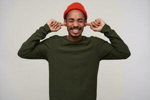 Studio photo of displeased young bearded dark skinned guy keeping eyes closed while covering his ears with index fingers because of loud sounds, isolated over white background