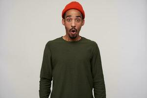 Overwhelmed young attractive bearded brunette male with dark skin looking amazedly at camera with round eyes, wearing red hat and khaki sweater over white background photo