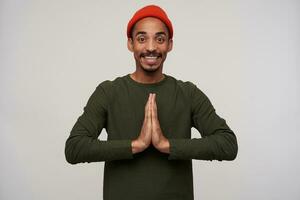 Positive young dark haired brown-eyed dark skinned man with beard folding raised palms together and looking happily at camera with wide smile, standing against white background photo