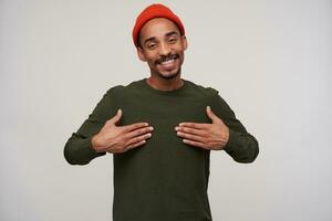 Pleased young dark haired bearded dark skinned guy holding palms on his chest and smiling happily to camera, being in nice mood while standing over white background photo