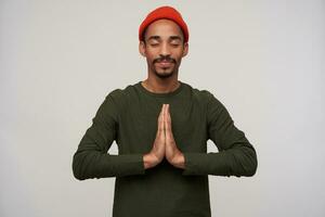 Pleasant looking young bearded dark skinned brunette guy raising hands in praying gesture while posing over white background, keeping eyes closed and smiling slightly photo