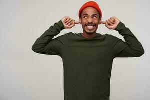 Disgruntled young brown-eyed dark skinned man with beard inserting forefingers into his ears while standing over white background, avoiding annoying sounds photo