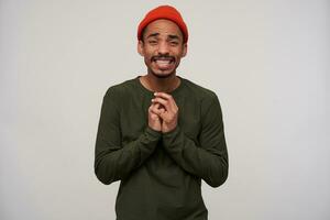 Portrait of young pretty bearded dark skinned brunette male folding hands in begging gesture and looking pityingly at camera, wearing casual clothes while standing over white background photo