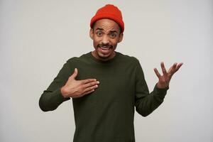 Excited young dark haired bearded male with dark skin raising palms and looking at camera with confused face, wearing red hat and khaki sweater while posing over white background photo