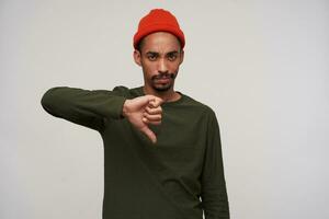 Portrait of dissatisfied young pretty bearded dark skinned man frowning eyebrows with folded lips and showing down with thumb, standing against white background in casual wear photo