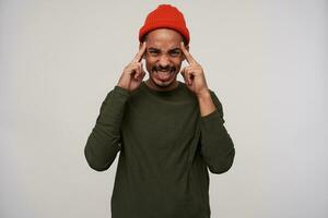 Dissatisfied young bearded dark skinned brunette guy looking painfully at camera and showing his perfect white teeth, holding forefingers on temples, standing against white background photo