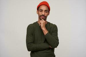 Thoughtful young bearded dark skinned brunette guy holding his chin and squinting while looking at camera, wearing casual clothes while posing over white background photo
