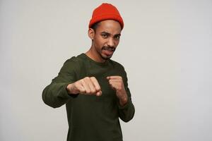 Handsome young brown-eyed dark skinned man with beard standing in defensive position with raised fists and looking wary, wearing red hat and khaki sweater over white background photo